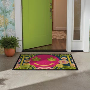 A rectangular doormat featuring a large hot pink smiley face on a deep teal background. The smiley face has green cannabis leaves for eyes and a yellow stitched-style smile. The central face is surrounded by stylized green foliage arches and concentric circles in pink, yellow, and white, resembling abstract flowers. The design mimics a vintage needlepoint or tapestry texture but is a flat print. The mat has a solid black trim border.