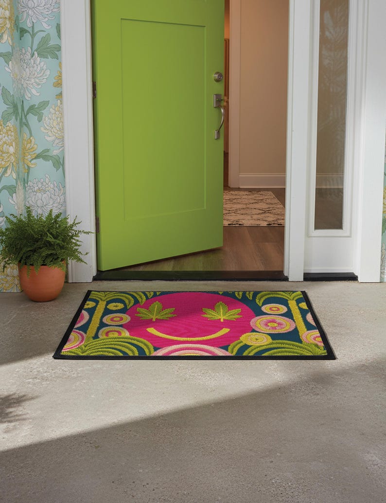 A rectangular doormat featuring a large hot pink smiley face on a deep teal background. The smiley face has green cannabis leaves for eyes and a yellow stitched-style smile. The central face is surrounded by stylized green foliage arches and concentric circles in pink, yellow, and white, resembling abstract flowers. The design mimics a vintage needlepoint or tapestry texture but is a flat print. The mat has a solid black trim border.