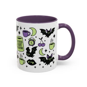White ceramic mug with vibrant purple interior and matching C-handle. Features a full wrap-around, 'spooky cute' pastel goth design. Central graphic is a lime green coffee cup with 'CERTIFIED NIGHT CREATURE' in black text. Surrounding illustrations include grumpy black bats, a steaming green coffee cup, a purple mug, vampire fangs, crescent moon, black cherries, and an alarm clock. Design uses bold graphics in black, lime green, and purple, accented with small black stars. Pastel Goth vibes.
