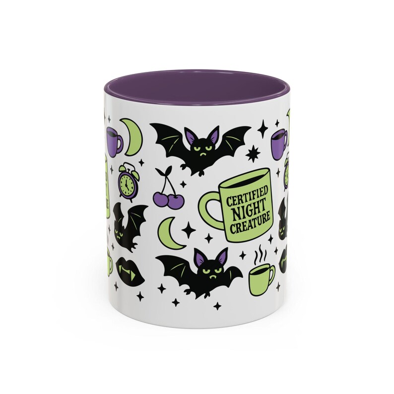 White ceramic mug with vibrant purple interior and matching C-handle. Features a full wrap-around, 'spooky cute' pastel goth design. Central graphic is a lime green coffee cup with 'CERTIFIED NIGHT CREATURE' in black text. Surrounding illustrations include grumpy black bats, a steaming green coffee cup, a purple mug, vampire fangs, crescent moon, black cherries, and an alarm clock. Design uses bold graphics in black, lime green, and purple, accented with small black stars. Pastel Goth vibes.
