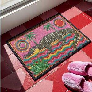 A rectangular doormat featuring a colorful 2D flat print designed to mimic raised, textured yarn embroidery. The central graphic showcases a large dark grey and yellow alligator resting in wavy water stripes of red, bright blue, and yellow. The background is hot pink, decorated with two green palm trees, a leafy green agave plant, and two stylized suns. The entire design is enclosed by a yellow border with repeating circle motifs and finished with a sleek black outer edge border trim.