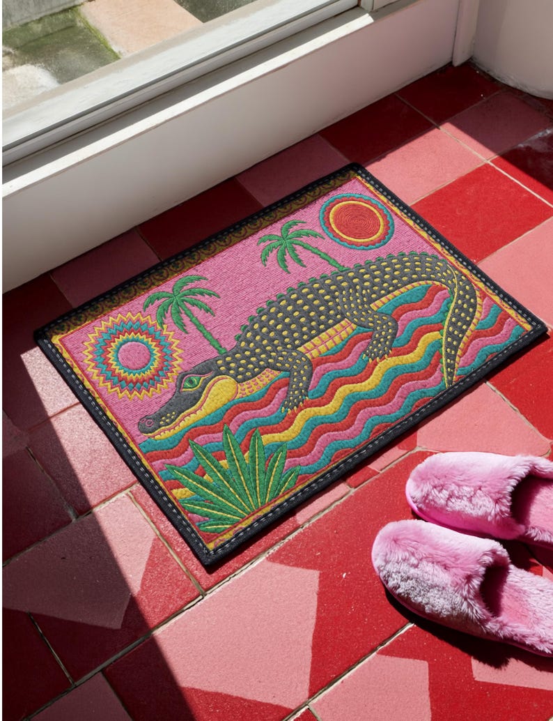 A rectangular doormat featuring a colorful 2D flat print designed to mimic raised, textured yarn embroidery. The central graphic showcases a large dark grey and yellow alligator resting in wavy water stripes of red, bright blue, and yellow. The background is hot pink, decorated with two green palm trees, a leafy green agave plant, and two stylized suns. The entire design is enclosed by a yellow border with repeating circle motifs and finished with a sleek black outer edge border trim.