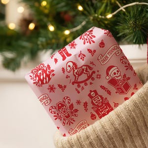 Premium satin finish wrapping paper featuring a repeating pastel goth pattern on a pink background. The design showcases a playful yet spooky aesthetic with high-contrast skeleton nutcrackers, skull jack-in-the-boxes, and rocking horse skeletons in soft pinks, and reds. The paper texture appears semi-glossy. Shown in three available sizes including sheets and rolls, specifically 29 inches wide, suitable for wrapping gifts for holidays, birthdays, or gothic themed occasions.