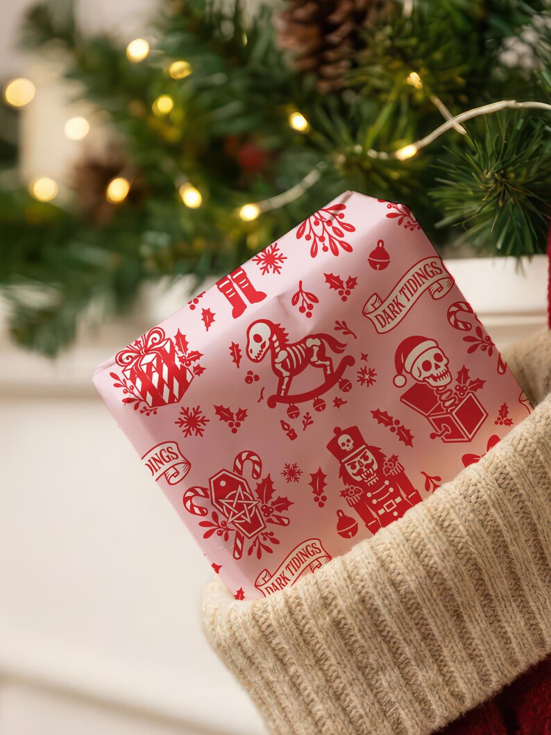 Premium satin finish wrapping paper featuring a repeating pastel goth pattern on a pink background. The design showcases a playful yet spooky aesthetic with high-contrast skeleton nutcrackers, skull jack-in-the-boxes, and rocking horse skeletons in soft pinks, and reds. The paper texture appears semi-glossy. Shown in three available sizes including sheets and rolls, specifically 29 inches wide, suitable for wrapping gifts for holidays, birthdays, or gothic themed occasions.