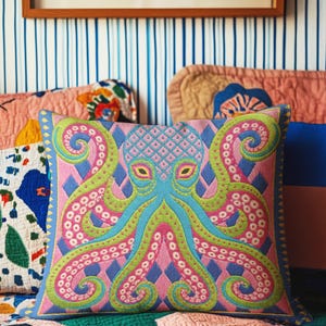 A square decorative throw pillow cover featuring a highly symmetrical bright octopus design. The octopus has a turquoise blue body with a diamond patterned head and eight curled tentacles detailed in bright pink and lime green. The background is a repeating diamond argyle pattern in royal blue and light pink. The artwork is a flat two dimensional print that mimics the textured look of intricate crewelwork embroidery. The pillow features a hidden zipper on the bottom edge. No insert included.