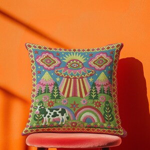 Description of a square throw pillow with a vibrant maximalist alien design. A central UFO with a large all-seeing eye hovers above a landscape with two rainbows, pink mountains, and green pine trees. A black and white spotted cow stands in the foreground. The design is a high-contrast palette of sky blue, magenta, hot pink, and meadow green. The pillow has a faux-embroidery 2D print finish on linen-feel fabric, framed by a yellow and pink scalloped floral border.