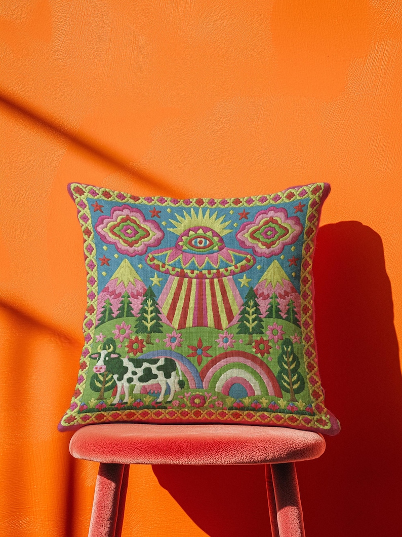 Description of a square throw pillow with a vibrant maximalist alien design. A central UFO with a large all-seeing eye hovers above a landscape with two rainbows, pink mountains, and green pine trees. A black and white spotted cow stands in the foreground. The design is a high-contrast palette of sky blue, magenta, hot pink, and meadow green. The pillow has a faux-embroidery 2D print finish on linen-feel fabric, framed by a yellow and pink scalloped floral border.