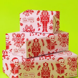 Premium satin finish wrapping paper featuring a repeating pastel goth pattern on a pink background. The design showcases a playful yet spooky aesthetic with high-contrast skeleton nutcrackers, skull jack-in-the-boxes, and rocking horse skeletons in soft pinks, and reds. The paper texture appears semi-glossy. Shown in three available sizes including sheets and rolls, specifically 29 inches wide, suitable for wrapping gifts for holidays, birthdays, or gothic themed occasions.