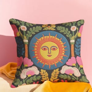 A square folk art sun throw pillow with a dark, charcoal-gray background. The central design is a vibrant celestial sun face in shades of orange, yellow, and red, with defined facial features. The sun is enclosed in a circular blue border with a textured, faux-embroidery pattern. Surrounding the central sun are symmetrical, colorful botanical and floral elements, including pink and white flowers, and green and gold leaves. The overall style is maximalist and bohemian.