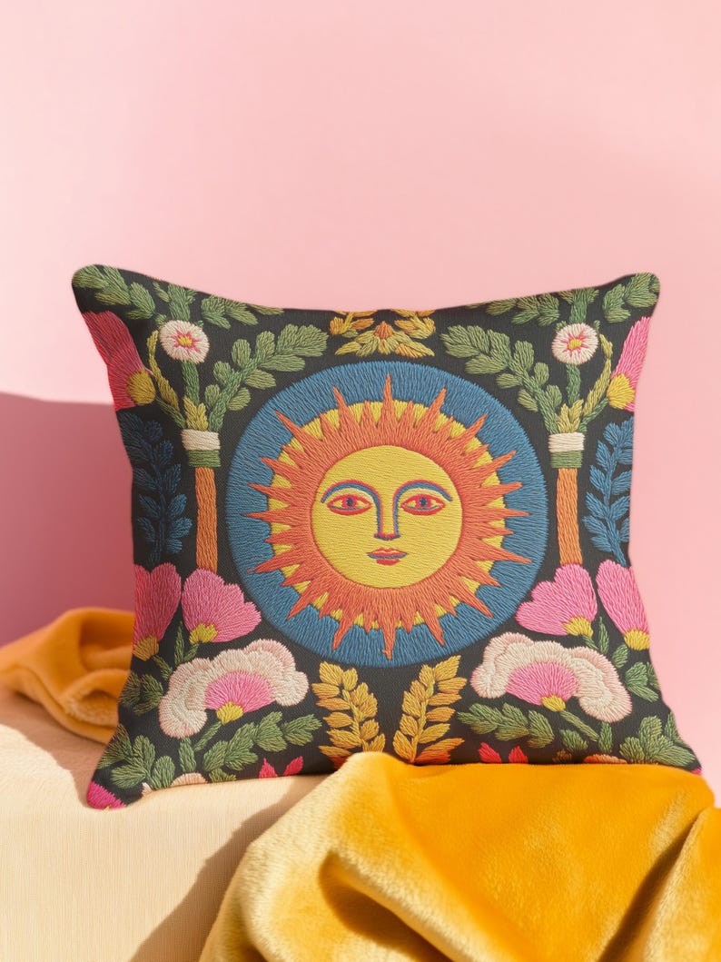 A square folk art sun throw pillow with a dark, charcoal-gray background. The central design is a vibrant celestial sun face in shades of orange, yellow, and red, with defined facial features. The sun is enclosed in a circular blue border with a textured, faux-embroidery pattern. Surrounding the central sun are symmetrical, colorful botanical and floral elements, including pink and white flowers, and green and gold leaves. The overall style is maximalist and bohemian.