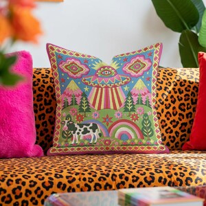 Description of a square throw pillow with a vibrant maximalist alien design. A central UFO with a large all-seeing eye hovers above a landscape with two rainbows, pink mountains, and green pine trees. A black and white spotted cow stands in the foreground. The design is a high-contrast palette of sky blue, magenta, hot pink, and meadow green. The pillow has a faux-embroidery 2D print finish on linen-feel fabric, framed by a yellow and pink scalloped floral border.