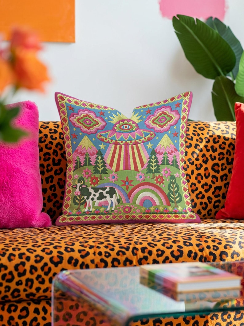 Description of a square throw pillow with a vibrant maximalist alien design. A central UFO with a large all-seeing eye hovers above a landscape with two rainbows, pink mountains, and green pine trees. A black and white spotted cow stands in the foreground. The design is a high-contrast palette of sky blue, magenta, hot pink, and meadow green. The pillow has a faux-embroidery 2D print finish on linen-feel fabric, framed by a yellow and pink scalloped floral border.