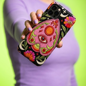 May include: Black phone case with colorful embroidered floral and celestial designs. The case features a pink planchette design with an eye, sun, and rainbow accents. The case has a black background with flowers, moons, and stars.