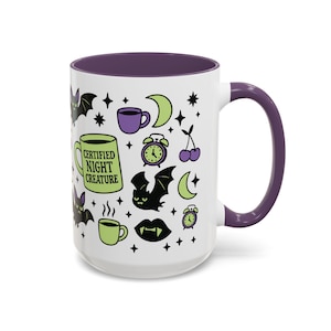 White ceramic mug with vibrant purple interior and matching C-handle. Features a full wrap-around, 'spooky cute' pastel goth design. Central graphic is a lime green coffee cup with 'CERTIFIED NIGHT CREATURE' in black text. Surrounding illustrations include grumpy black bats, a steaming green coffee cup, a purple mug, vampire fangs, crescent moon, black cherries, and an alarm clock. Design uses bold graphics in black, lime green, and purple, accented with small black stars. Pastel Goth vibes.