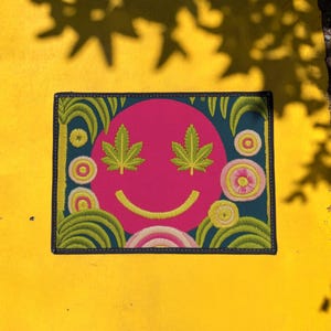 A rectangular doormat featuring a large hot pink smiley face on a deep teal background. The smiley face has green cannabis leaves for eyes and a yellow stitched-style smile. The central face is surrounded by stylized green foliage arches and concentric circles in pink, yellow, and white, resembling abstract flowers. The design mimics a vintage needlepoint or tapestry texture but is a flat print. The mat has a solid black trim border.