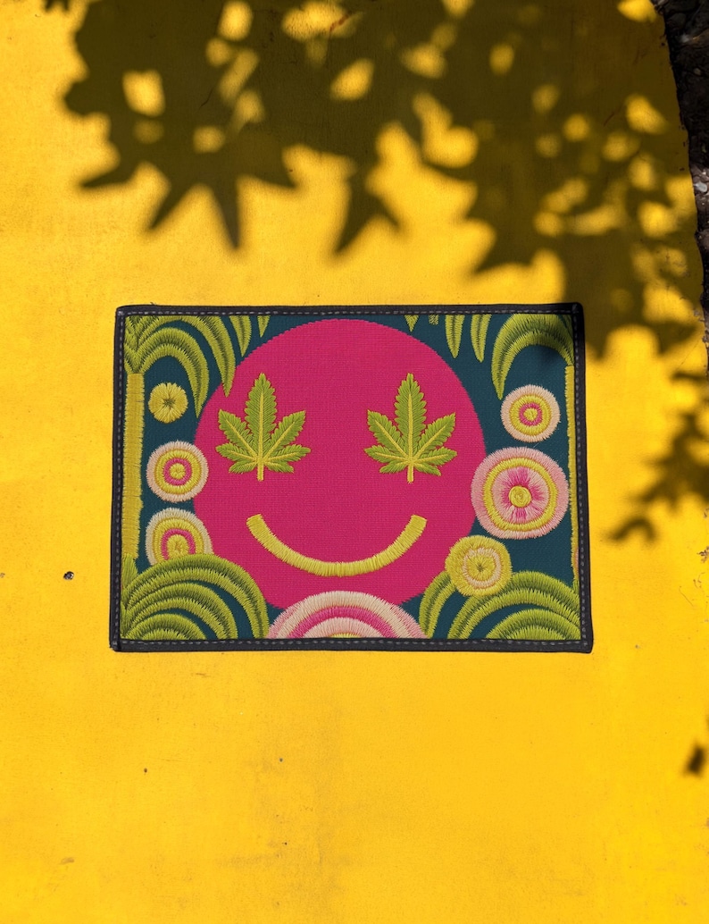 A rectangular doormat featuring a large hot pink smiley face on a deep teal background. The smiley face has green cannabis leaves for eyes and a yellow stitched-style smile. The central face is surrounded by stylized green foliage arches and concentric circles in pink, yellow, and white, resembling abstract flowers. The design mimics a vintage needlepoint or tapestry texture but is a flat print. The mat has a solid black trim border.