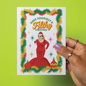 May include: A Christmas greeting card with the text "Have Yourself a Filthy Christmas." The card features a cartoon illustration of a person in a red dress and Santa hat, holding a camera. The card is decorated with Christmas ornaments and garland.