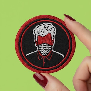 Peut inclure: Écusson brodé rond avec un motif de visage stylisé. Le visage a des cheveux blancs, un masque rouge et une chemise noire. Le motif est sur fond noir avec un bord rouge. Sur fond vert.