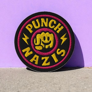 May include: Embroidered patch with the words "PUNCH NAZIS" in yellow, arched around a black circle. The patch features a pink border and a yellow fist graphic in the center, with yellow lightning bolts.
