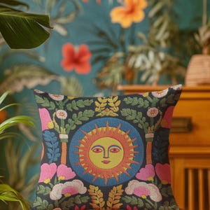 A square folk art sun throw pillow with a dark, charcoal-gray background. The central design is a vibrant celestial sun face in shades of orange, yellow, and red, with defined facial features. The sun is enclosed in a circular blue border with a textured, faux-embroidery pattern. Surrounding the central sun are symmetrical, colorful botanical and floral elements, including pink and white flowers, and green and gold leaves. The overall style is maximalist and bohemian.