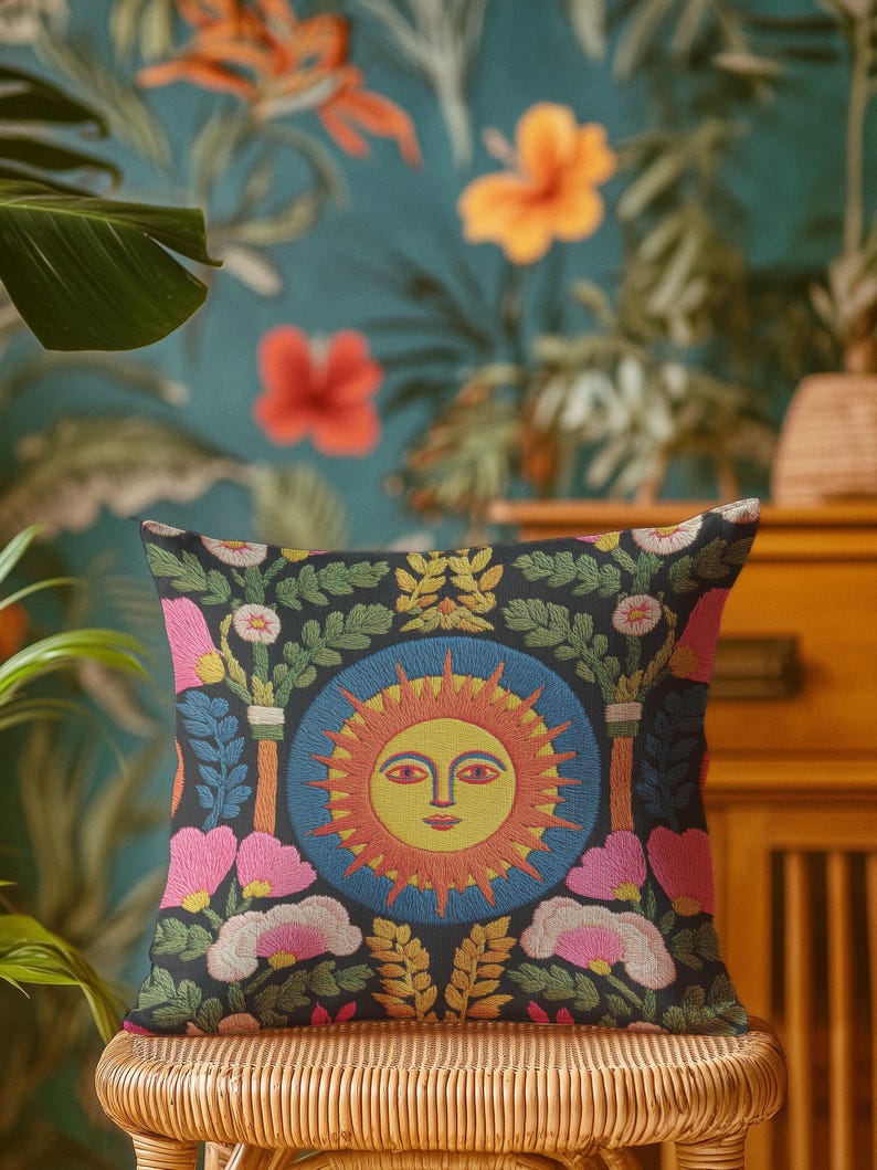 A square folk art sun throw pillow with a dark, charcoal-gray background. The central design is a vibrant celestial sun face in shades of orange, yellow, and red, with defined facial features. The sun is enclosed in a circular blue border with a textured, faux-embroidery pattern. Surrounding the central sun are symmetrical, colorful botanical and floral elements, including pink and white flowers, and green and gold leaves. The overall style is maximalist and bohemian.