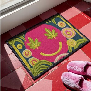 A rectangular doormat featuring a large hot pink smiley face on a deep teal background. The smiley face has green cannabis leaves for eyes and a yellow stitched-style smile. The central face is surrounded by stylized green foliage arches and concentric circles in pink, yellow, and white, resembling abstract flowers. The design mimics a vintage needlepoint or tapestry texture but is a flat print. The mat has a solid black trim border.
