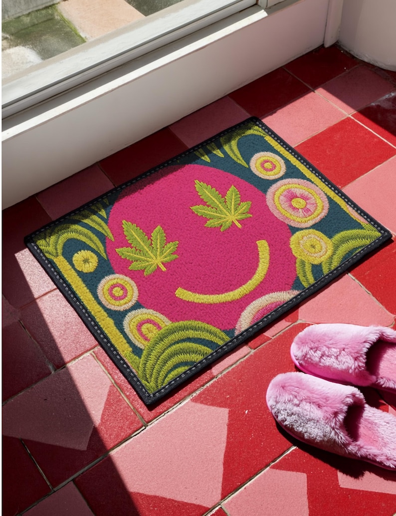 A rectangular doormat featuring a large hot pink smiley face on a deep teal background. The smiley face has green cannabis leaves for eyes and a yellow stitched-style smile. The central face is surrounded by stylized green foliage arches and concentric circles in pink, yellow, and white, resembling abstract flowers. The design mimics a vintage needlepoint or tapestry texture but is a flat print. The mat has a solid black trim border.