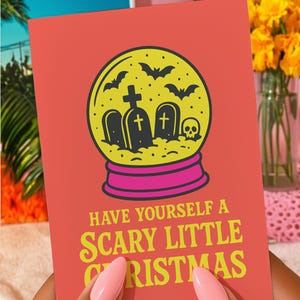 A vibrant, flat-lay photograph of the "Scary Little Christmas" greeting card. The card has a bright orange background with bold, retro yellow text. The central graphic is a snow globe with a yellow glass dome and a hot pink base. Inside the globe is a black silhouette of a spooky graveyard scene, featuring tombstones with crosses, a skull, and a swarm of flying bats. The card is made from premium 120lb silk cover stock with a slight sheen and measures 5x7 inches.