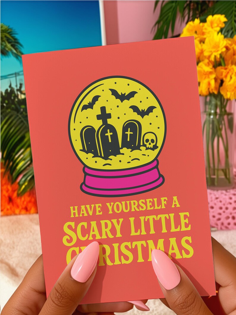 A vibrant, flat-lay photograph of the "Scary Little Christmas" greeting card. The card has a bright orange background with bold, retro yellow text. The central graphic is a snow globe with a yellow glass dome and a hot pink base. Inside the globe is a black silhouette of a spooky graveyard scene, featuring tombstones with crosses, a skull, and a swarm of flying bats. The card is made from premium 120lb silk cover stock with a slight sheen and measures 5x7 inches.