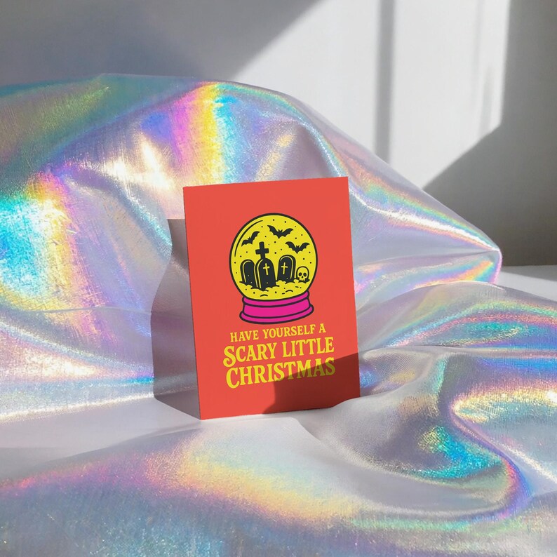 A vibrant, flat-lay photograph of the "Scary Little Christmas" greeting card. The card has a bright orange background with bold, retro yellow text. The central graphic is a snow globe with a yellow glass dome and a hot pink base. Inside the globe is a black silhouette of a spooky graveyard scene, featuring tombstones with crosses, a skull, and a swarm of flying bats. The card is made from premium 120lb silk cover stock with a slight sheen and measures 5x7 inches.