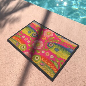 Rectangular indoor outdoor doormat featuring a vibrant hot pink background with a symmetrical maximalist pattern. The design showcases stylized sardines in yellow, lime green, and cobalt blue swimming horizontally towards a central vertical axis of blooming pale pink flowers and green vines. The imagery mimics the texture of vintage needlepoint embroidery but is a flat print on polyester. The mat is framed by a sleek black trim and features a non-slip rubber backing for safety.