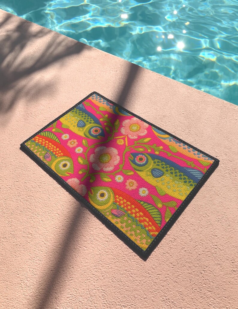 Rectangular indoor outdoor doormat featuring a vibrant hot pink background with a symmetrical maximalist pattern. The design showcases stylized sardines in yellow, lime green, and cobalt blue swimming horizontally towards a central vertical axis of blooming pale pink flowers and green vines. The imagery mimics the texture of vintage needlepoint embroidery but is a flat print on polyester. The mat is framed by a sleek black trim and features a non-slip rubber backing for safety.