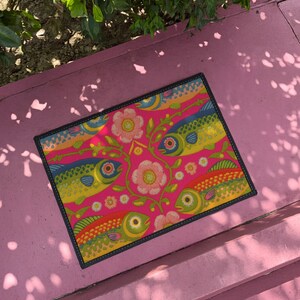 Rectangular indoor outdoor doormat featuring a vibrant hot pink background with a symmetrical maximalist pattern. The design showcases stylized sardines in yellow, lime green, and cobalt blue swimming horizontally towards a central vertical axis of blooming pale pink flowers and green vines. The imagery mimics the texture of vintage needlepoint embroidery but is a flat print on polyester. The mat is framed by a sleek black trim and features a non-slip rubber backing for safety.