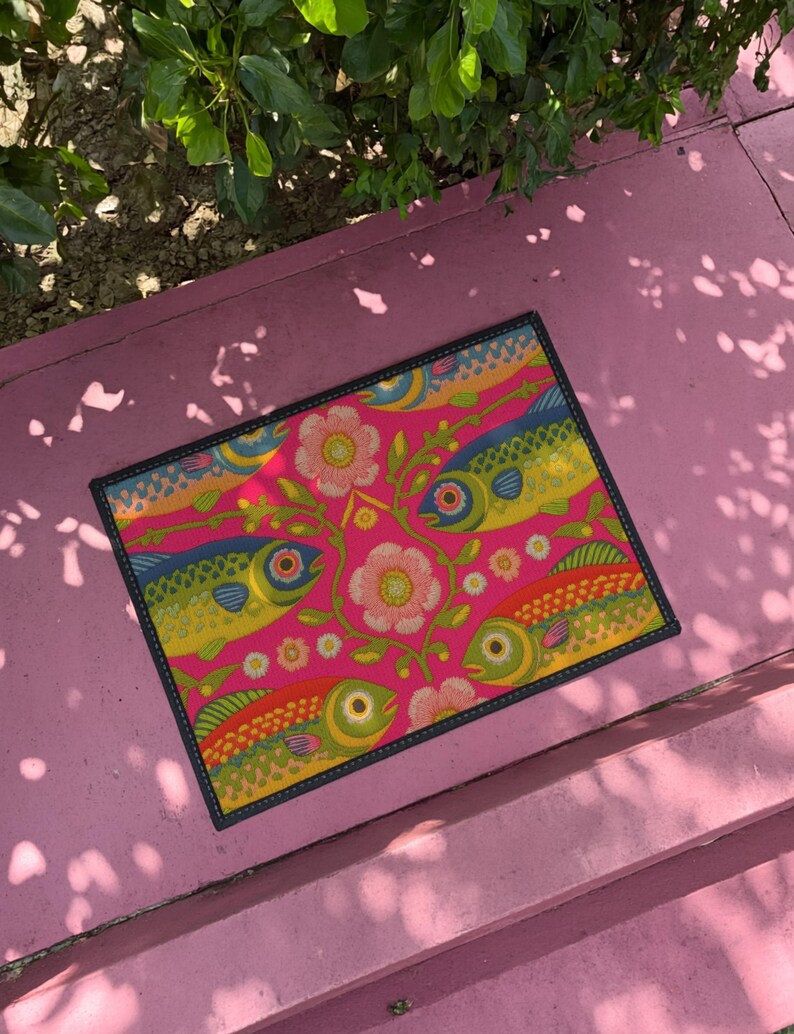 Rectangular indoor outdoor doormat featuring a vibrant hot pink background with a symmetrical maximalist pattern. The design showcases stylized sardines in yellow, lime green, and cobalt blue swimming horizontally towards a central vertical axis of blooming pale pink flowers and green vines. The imagery mimics the texture of vintage needlepoint embroidery but is a flat print on polyester. The mat is framed by a sleek black trim and features a non-slip rubber backing for safety.