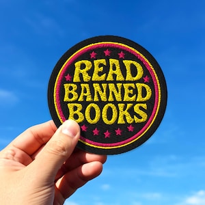 May include: A round black patch with the words "READ BANNED BOOKS" embroidered in yellow, outlined in pink and yellow. Pink stars are embroidered around the edge. The patch is held against a blue sky.
