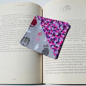 May include: Two fabric bookmarks with a cat and a bird design on a gray background. The bookmarks are folded in a triangle shape and are made of a pink and purple fabric with a polka dot pattern.