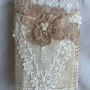 Op de afbeelding: Een beige stoffen journalhoes met delicate witte kant en een grote beige bloemenapplicatie.