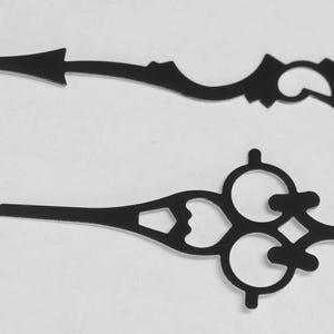 May include: Two black metal clock hands with intricate designs. The longer hand has a circular end and the shorter hand has a three-circle design.