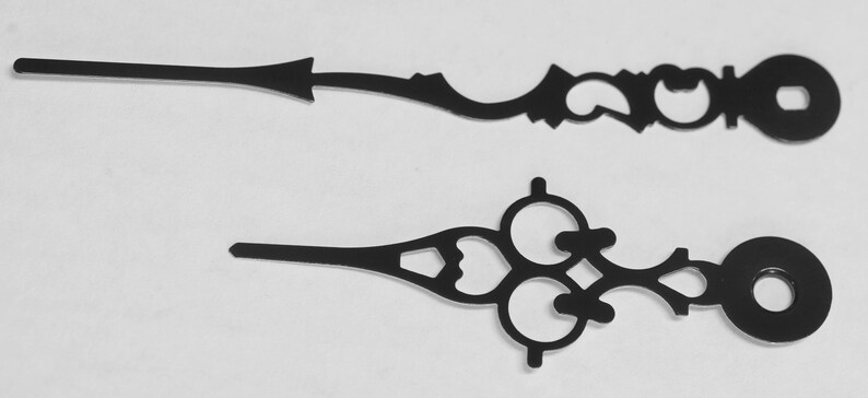May include: Two black metal clock hands with intricate designs. The longer hand has a circular end and the shorter hand has a three-circle design.
