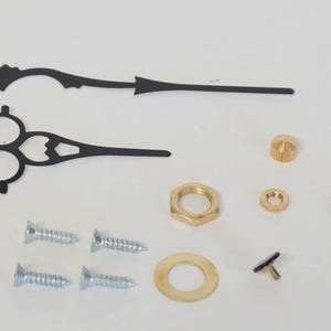 May include: A set of black clock hands with decorative details, including two screws, a washer, a nut, a small gold knob, a gold washer, and a black clock face insert.
