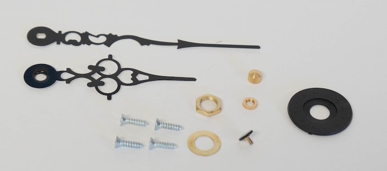 May include: A set of black clock hands with decorative details, including two screws, a washer, a nut, a small gold knob, a gold washer, and a black clock face insert.