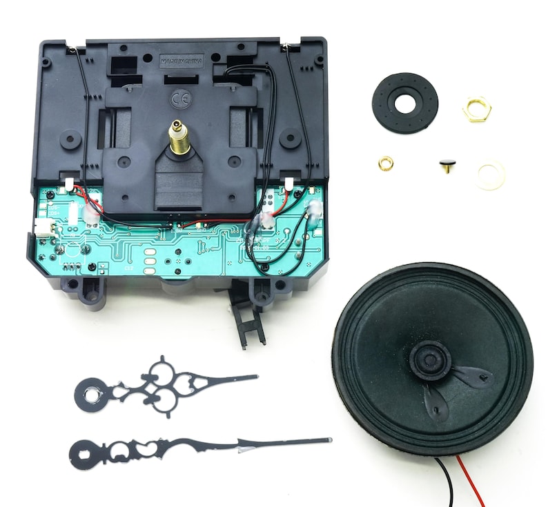 May include: Clock movement kit with black hands, a speaker, and a circuit board. The kit includes a black plastic clock movement with a green circuit board, a black speaker, and two black clock hands. The kit also includes a small black plastic washer, a small gold metal washer, and a small gold metal nut.