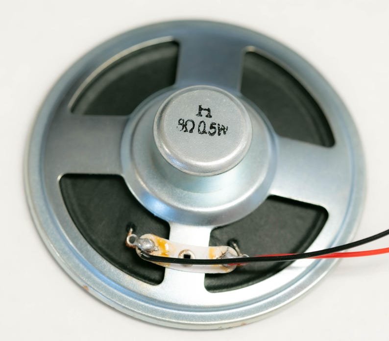 May include: A silver speaker with a black speaker cone and a silver dome with the text "8&Omega; 0.25W" printed on it. The speaker has two wires attached to it, one red and one black.