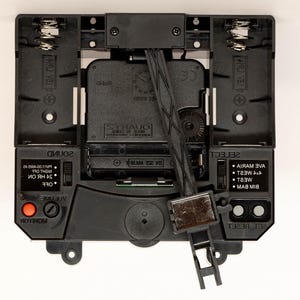 May include: Close-up of a black clock mechanism with a quartz movement. The clock has a volume control, a monitor button, a sound switch, and a select button with options for different chime melodies. The clock also has a reset button and a battery compartment.