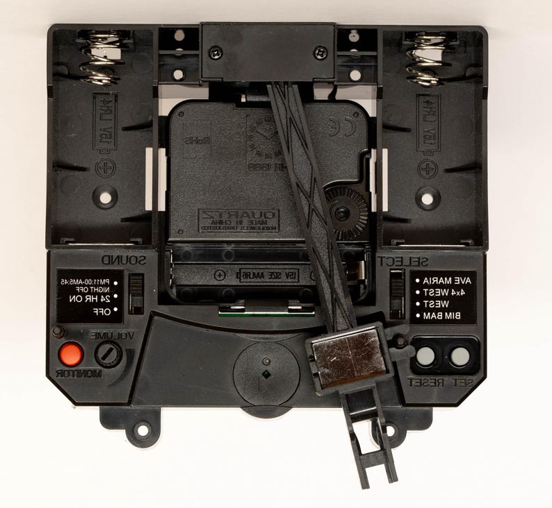May include: Close-up of a black clock mechanism with a quartz movement. The clock has a volume control, a monitor button, a sound switch, and a select button with options for different chime melodies. The clock also has a reset button and a battery compartment.