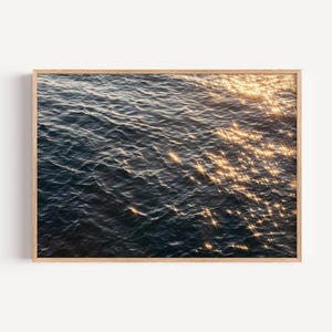 Puede incluir: Una impresión enmarcada que presenta una vista de cerca del agua del océano. El agua es de un azul profundo, con pequeñas olas y ondulaciones. La luz del sol se refleja en la superficie, creando reflejos brillantes. El marco es de madera clara.