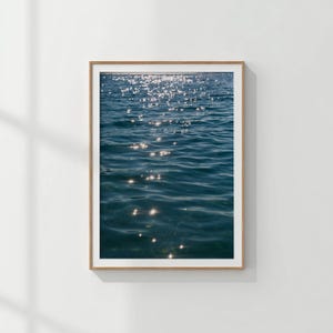 Puede incluir: Una impresión enmarcada que presenta un primer plano del agua del océano. El agua es de color azul profundo, con reflejos brillantes de la luz solar. La impresión está en un marco de madera marrón claro con un borde blanco.
