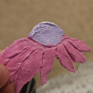 Puede incluir: Una flor de papel hecha a mano con una superficie texturizada. Los pétalos son de un rosa vibrante y el centro es de un color lavanda contrastante. La flor es parcialmente visible, lo que sugiere un artículo artesanal o decorativo.
