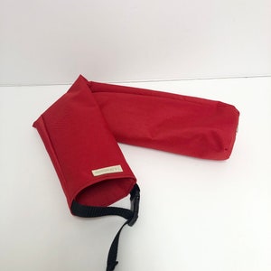 Pu&ograve; includere: Una custodia in tessuto rosso di forma cilindrica con una tracolla nera regolabile dotata di fibbia in plastica. Una piccola etichetta rettangolare con il testo J.A.S.Fashions &egrave; cucita sull'esterno del materiale tessile rosso.