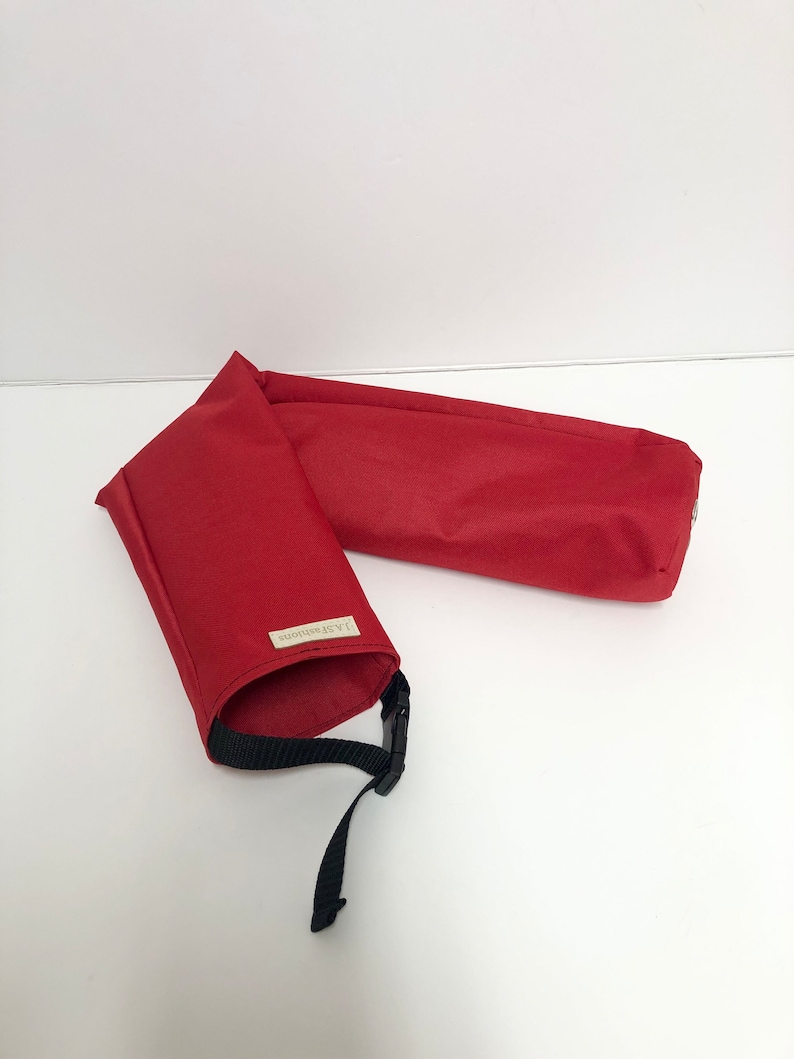 Pu&ograve; includere: Una custodia in tessuto rosso di forma cilindrica con una tracolla nera regolabile dotata di fibbia in plastica. Una piccola etichetta rettangolare con il testo J.A.S.Fashions &egrave; cucita sull'esterno del materiale tessile rosso.